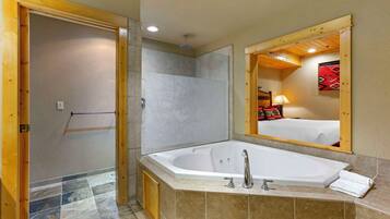 Un baño con una bañera grande, zona de ducha y un espejo que refleja una cama con una colcha roja.