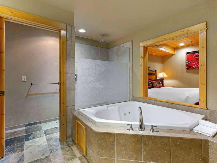 Un baño con una bañera grande, zona de ducha y un espejo que refleja una cama con una colcha roja.