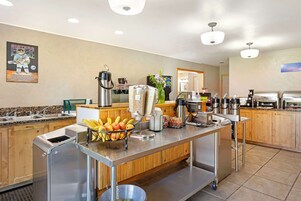 Zona de desayuno autoservicio con fruteros, máquinas de café y una barra de buffet.