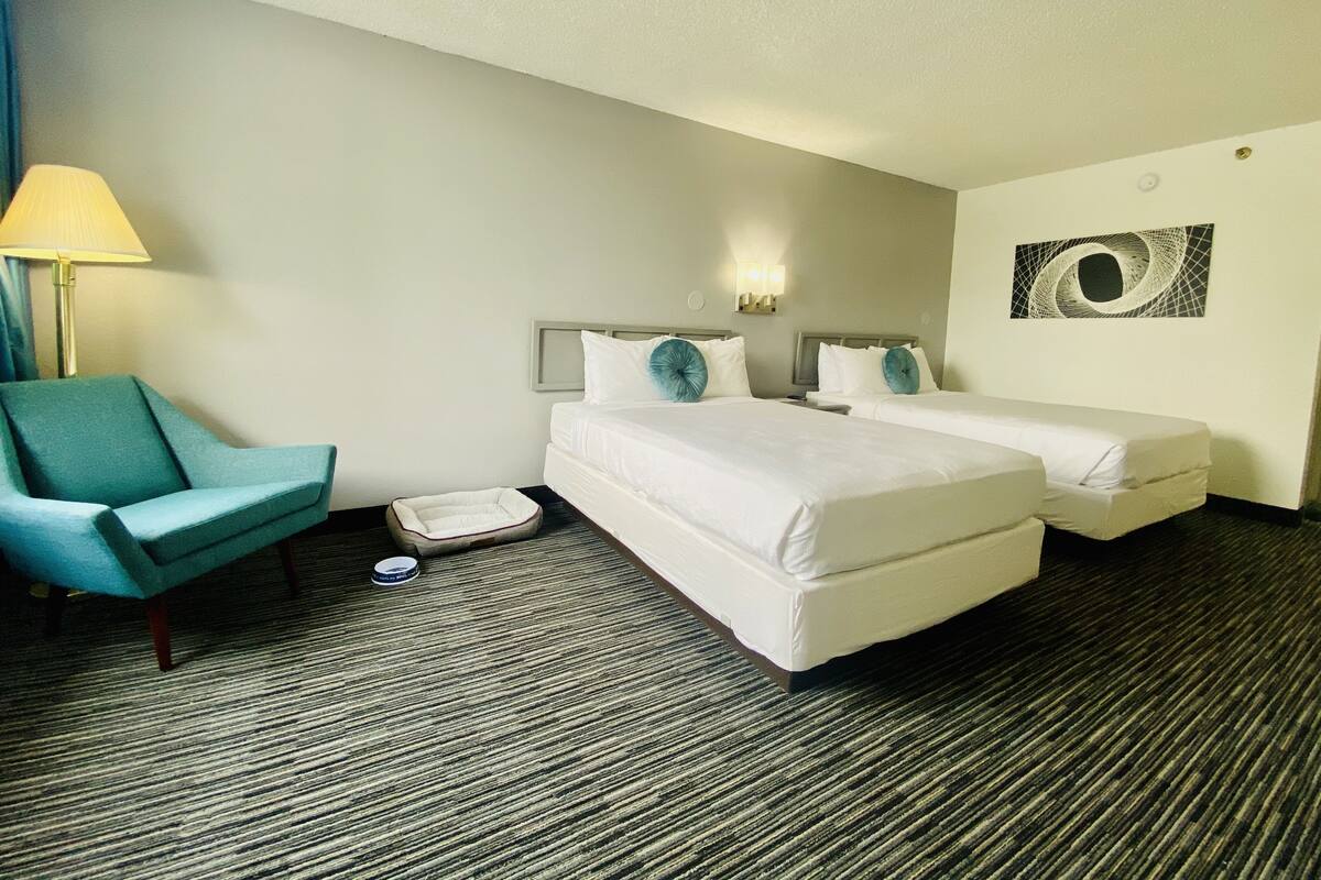 Una habitación de hotel con dos camas, una silla, una lámpara y una obra de arte en la pared.