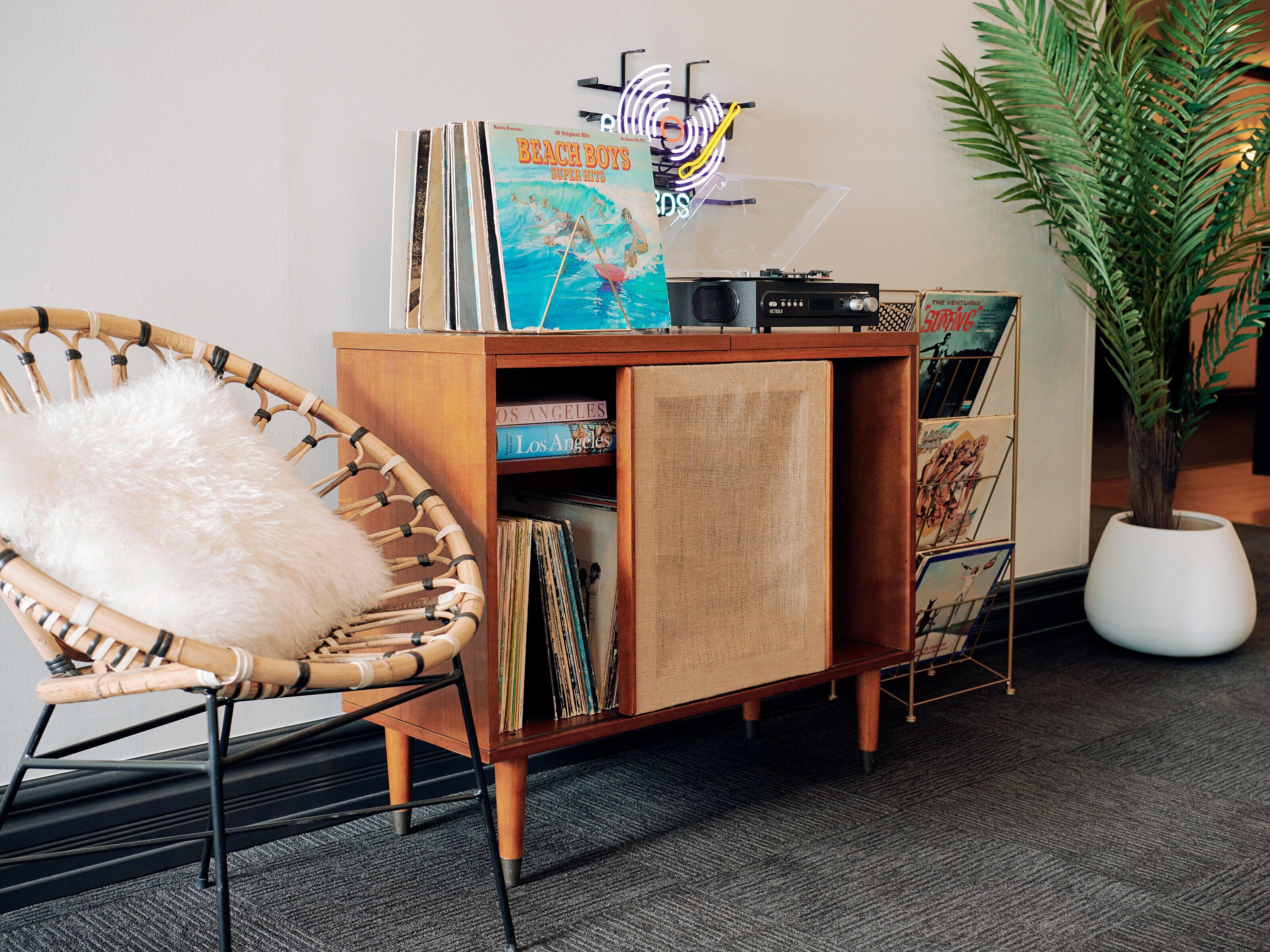 A room with a wooden cabinet displaying vinyl records, a wicker chair with a fluffy pillow, and a potted plant.