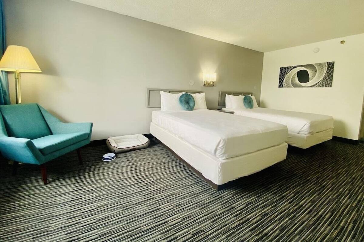 Habitación de hotel con dos camas, un sillón azul, una lámpara y una obra de arte en la pared.