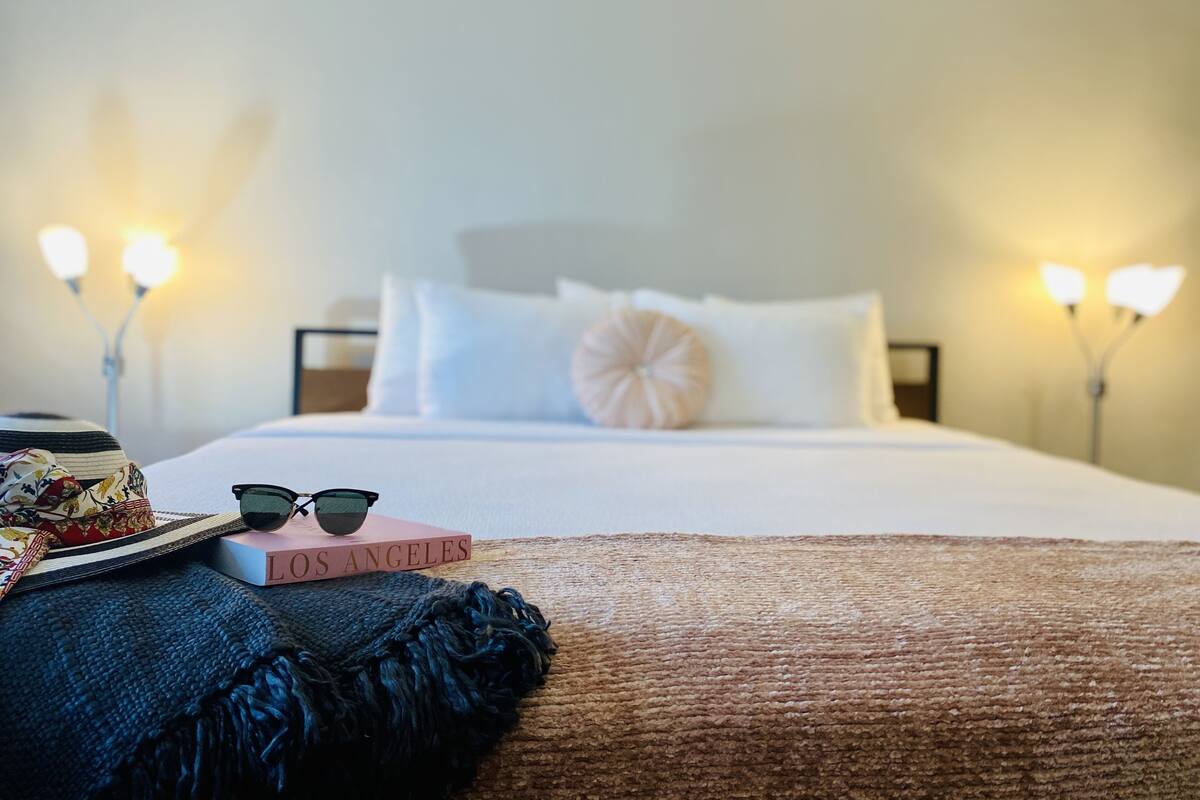 Una cama bien hecha con cubrecama y almohadas blancas, una mesita de noche con un sombrero, gafas de sol y un libro titulado “LOS ANGELES”, y dos lámparas de noche.