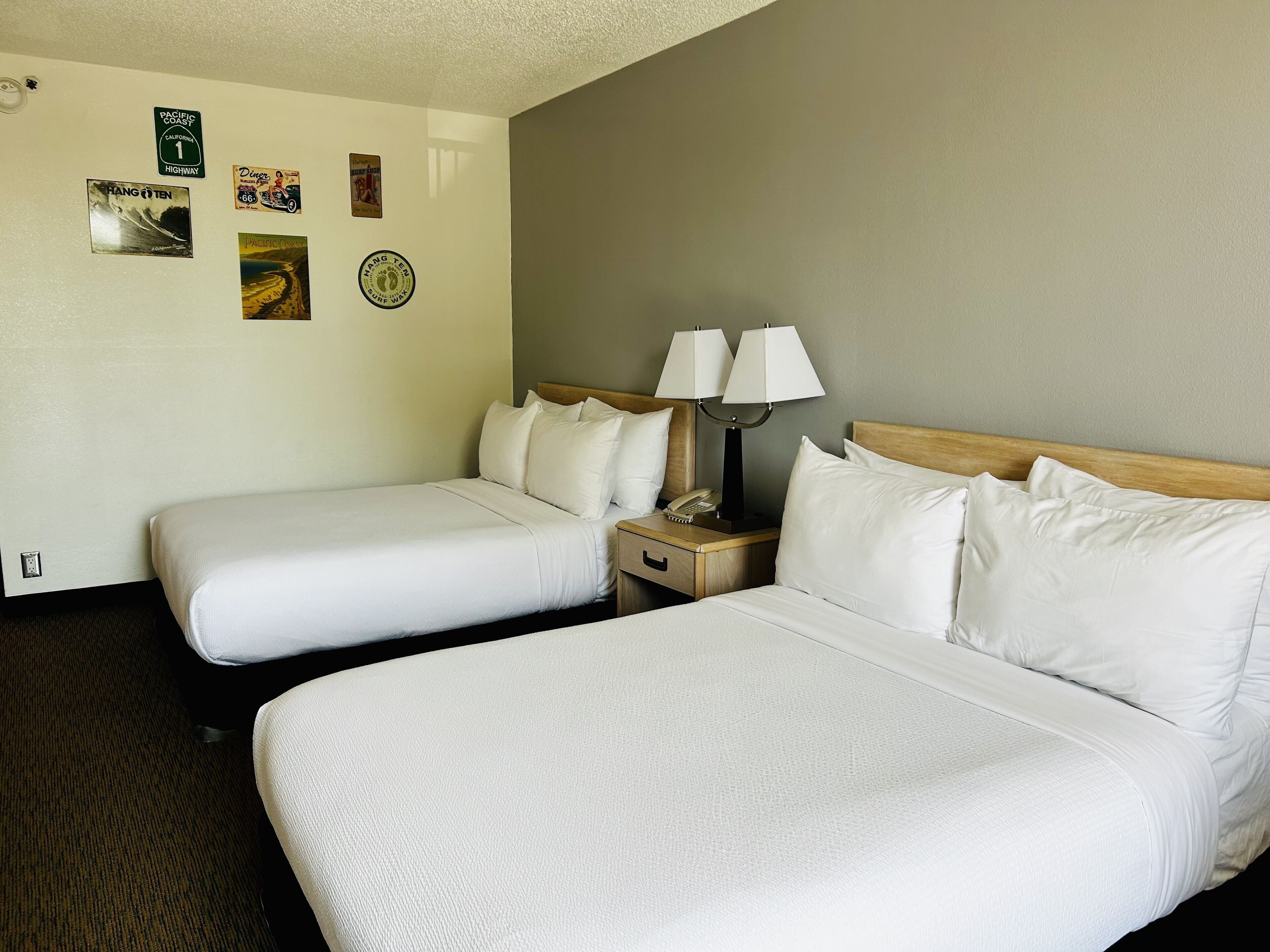 A hotel room with two beds, a nightstand with a lamp, and wall decorations including a 'Pacific Coast Highway' sign and a 'Hang Ten' surfboard.