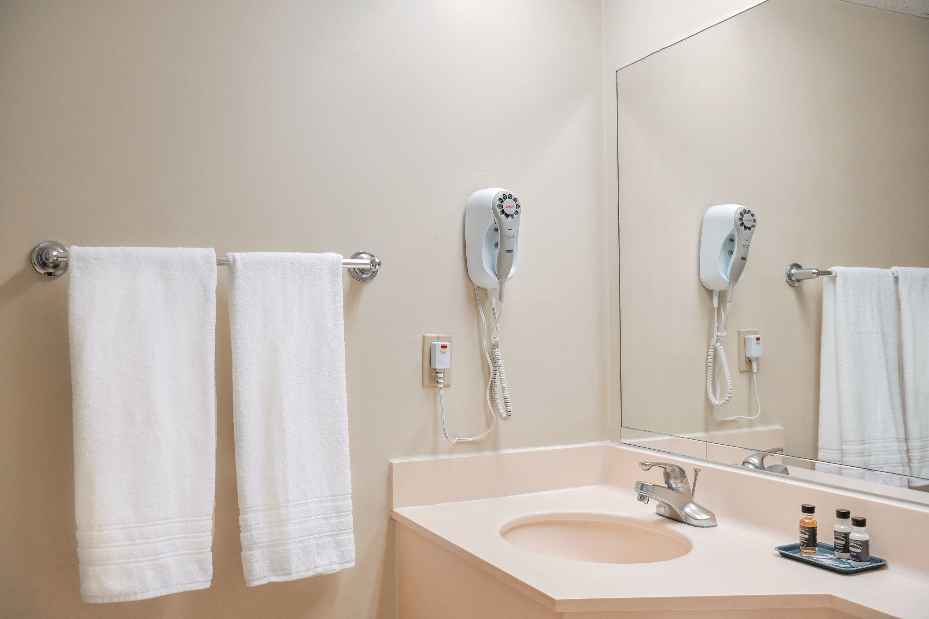 A bathroom with two white towels hanging on a towel bar, a hairdryer mounted on the wall, a sink with a mirror, and a small tray with toiletries.