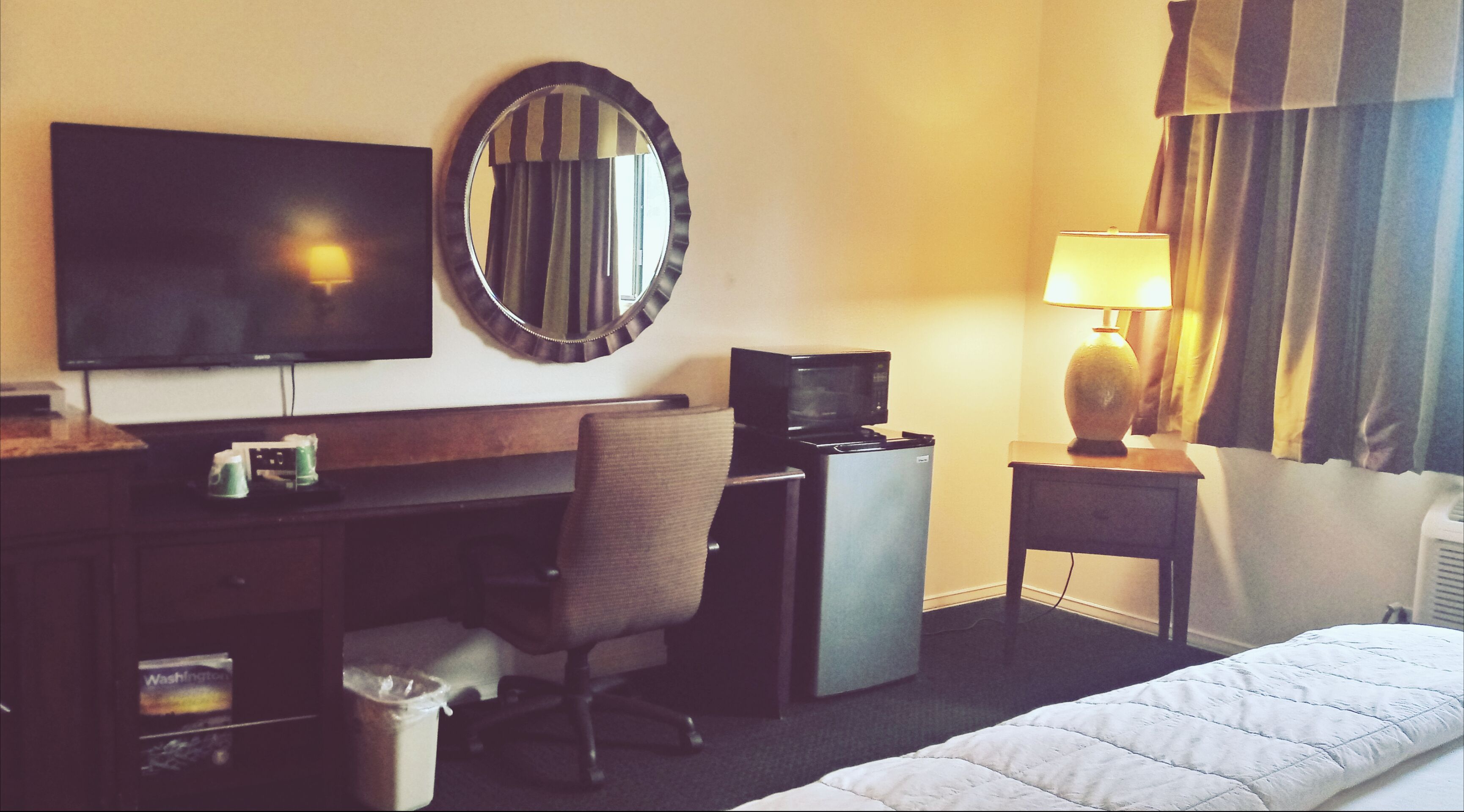 Una habitación de hotel con cama, escritorio con computadora, televisión, espejo, lámpara y basurero.