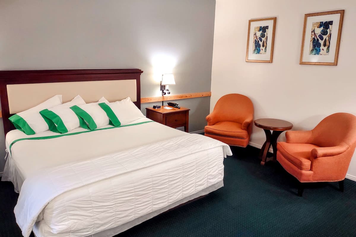 A hotel room with a large bed, two orange armchairs, a small table, and a nightstand with a lamp.
