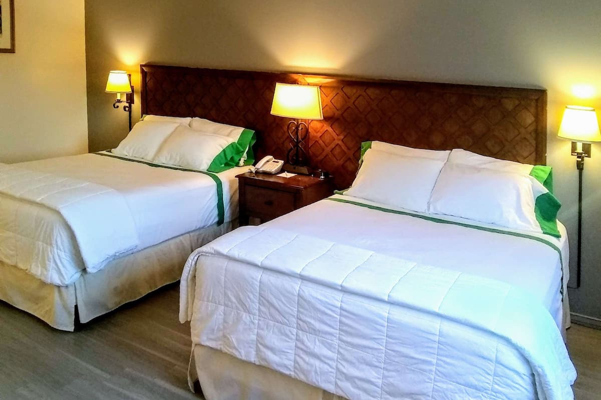 A hotel room with two single beds, each with white bedding and green accents. There are bedside lamps on either side of the beds, and a wooden headboard above them.