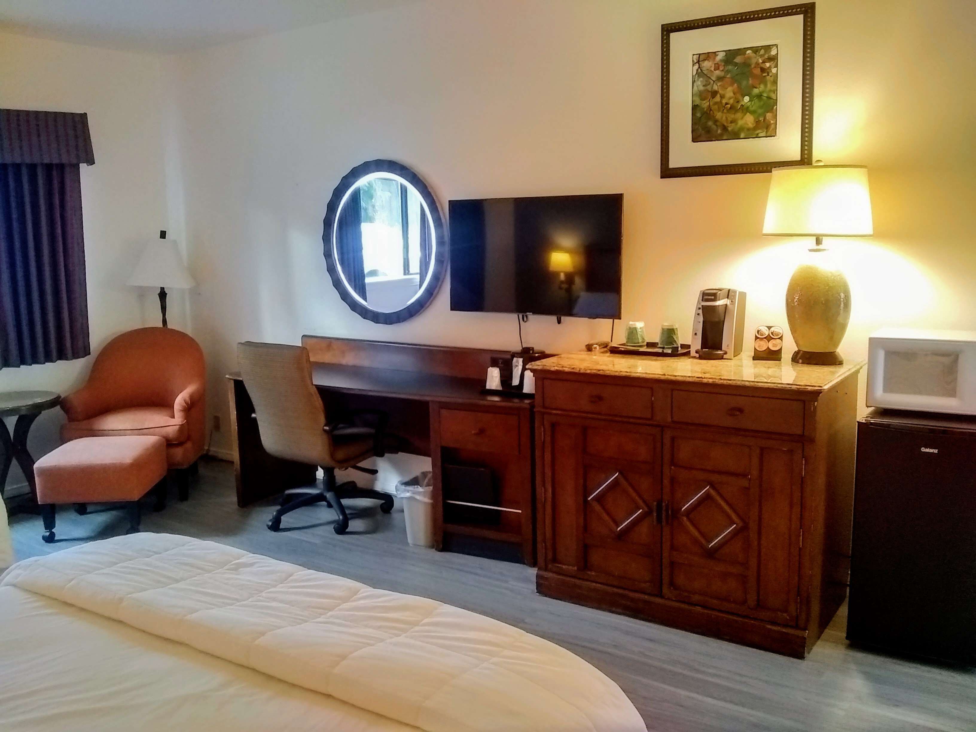 Quarto de hotel com cama, escrivaninha com cadeira, televisão, luminária, cafeteira e micro-ondas.