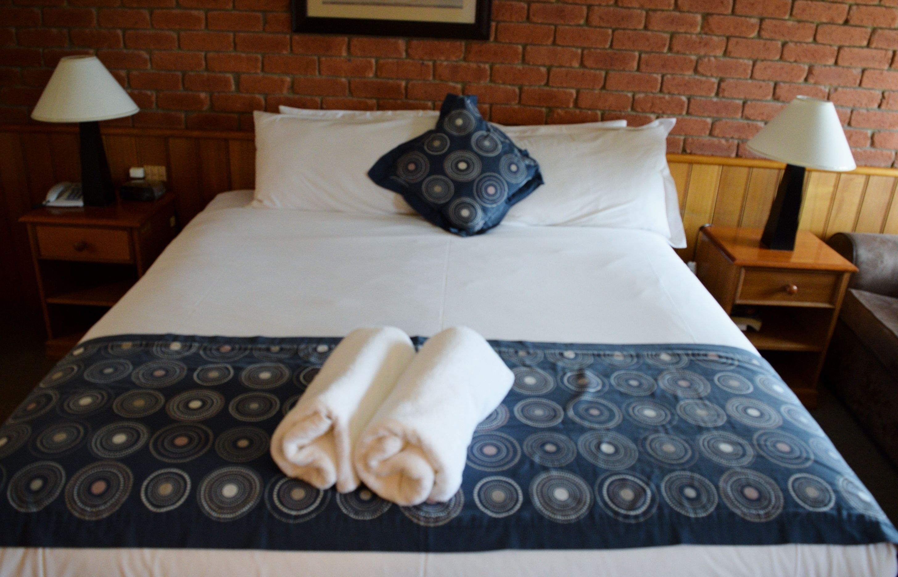 A neatly made bed with a patterned bedspread, two pillows, and folded towels on a wooden nightstand.