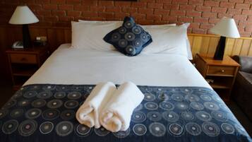 A neatly made bed with a patterned bedspread, two pillows, and folded towels on a wooden nightstand.