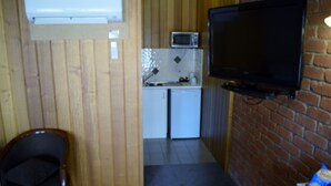 Una habitación compacta con un separador de madera, una pequeña cocina con microondas, un televisor fijado a una pared de ladrillo, una silla de madera y una mesita con aperitivos.