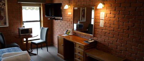 A hotel room with a brick wall, a bed with a patterned bedspread, a desk with a computer, a chair, a television mounted on the wall, and a small table with a lamp.