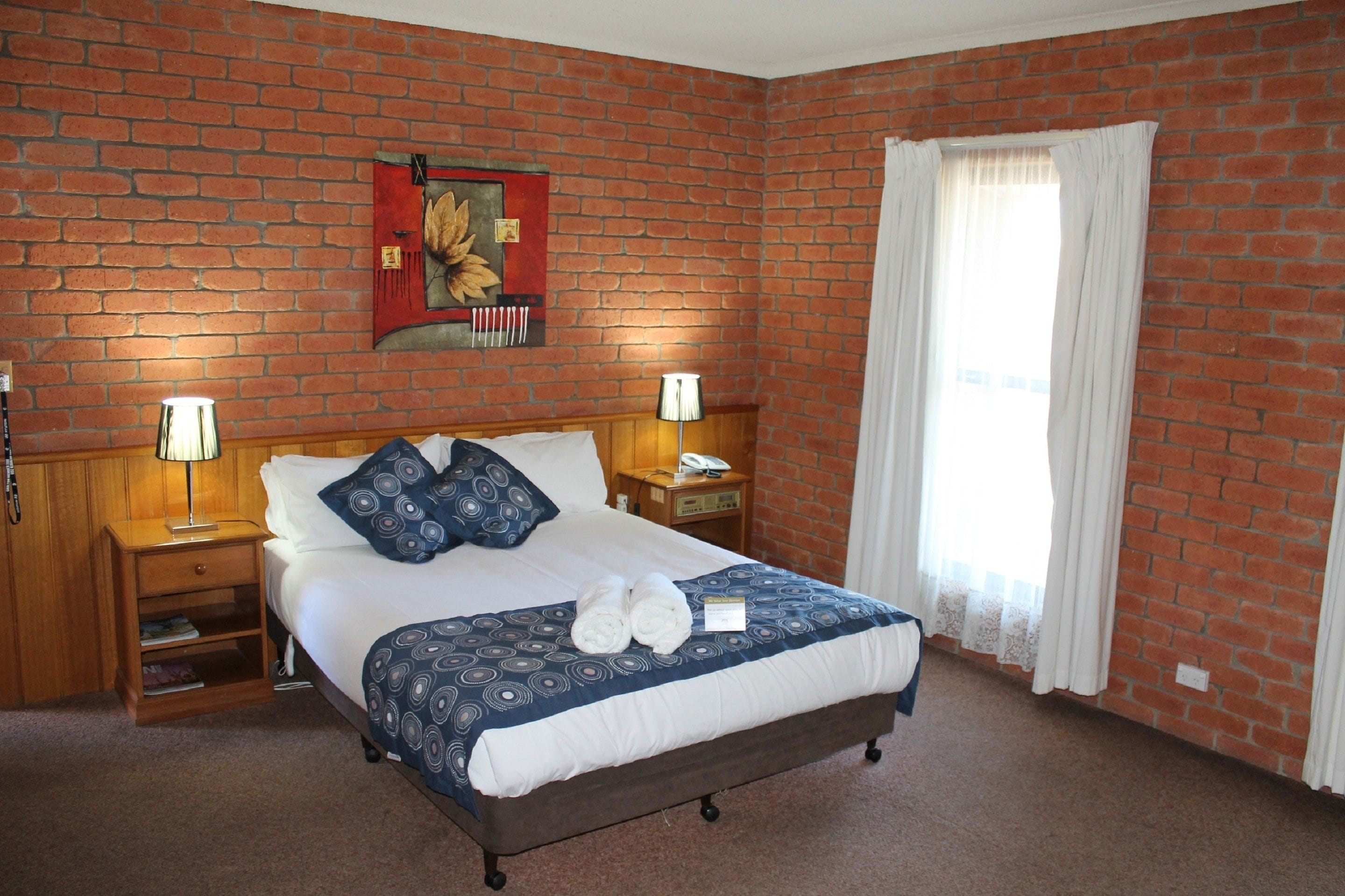 Une chambre d’hôtel avec un mur en briques, un lit recouvert d’une couvre-lit à motifs, deux lampes de chevet, une table de chevet, un téléphone et une fenêtre avec des rideaux.