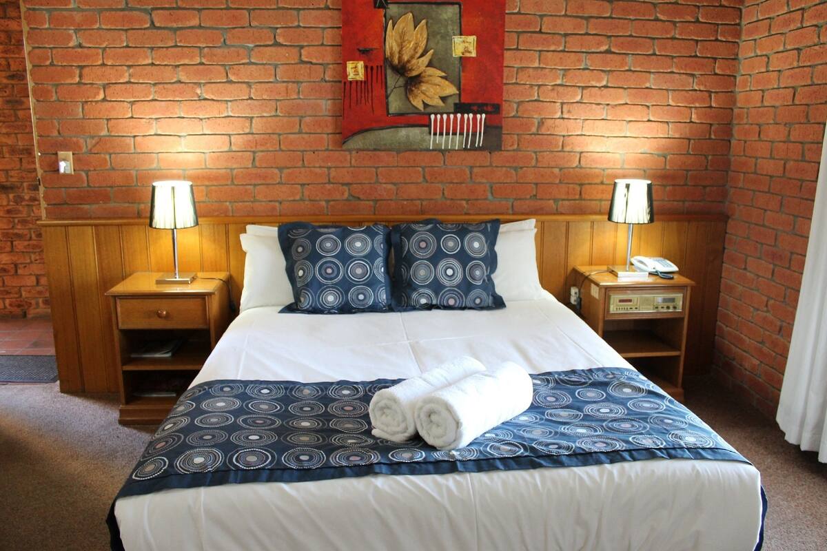 A hotel room with a brick wall, a bed with a blue and white patterned bedspread, two bedside lamps, a wooden nightstand, and a red artwork above the bed.