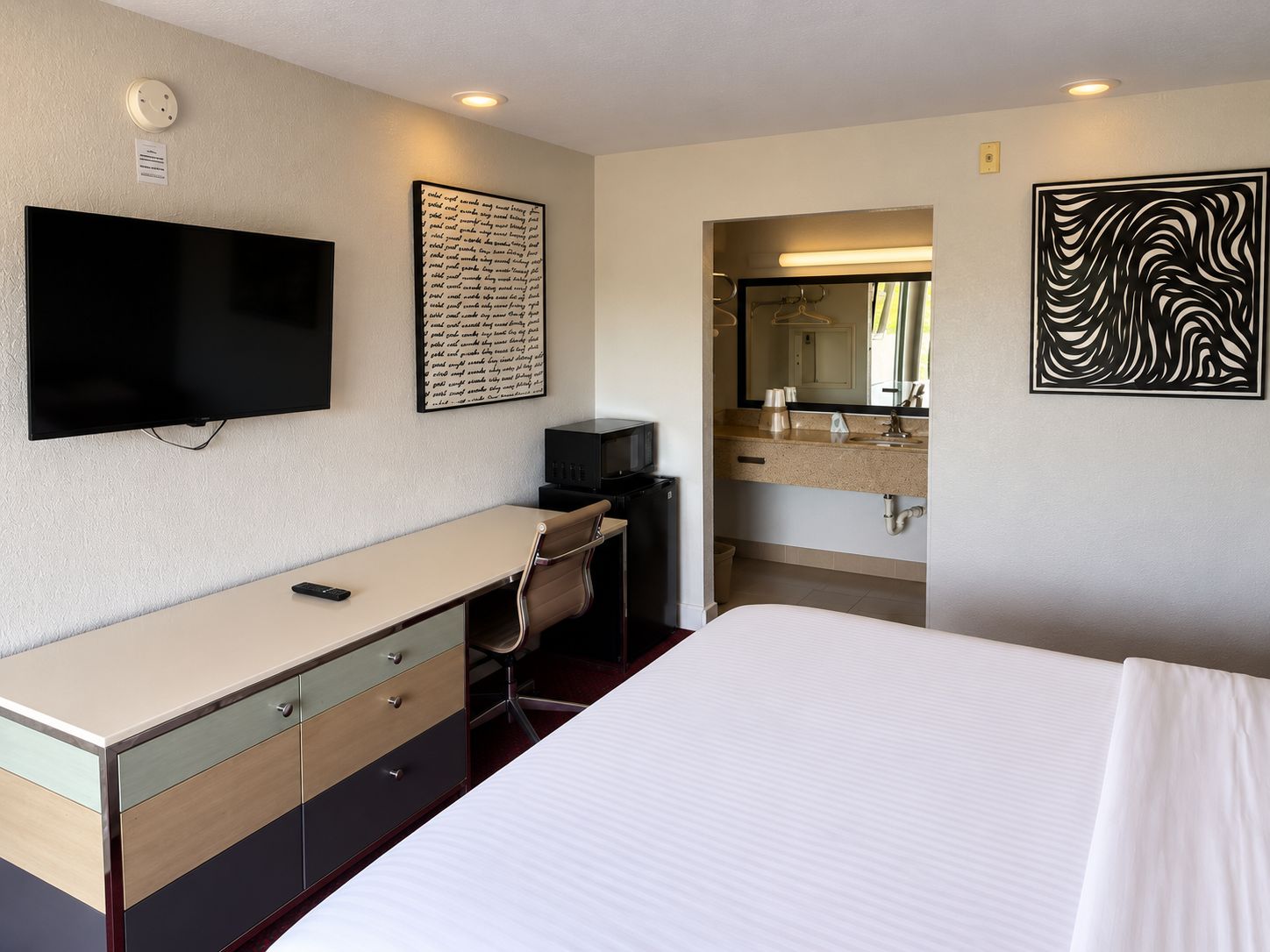 A hotel room with a bed, a desk with drawers, a television, a mirror, and artwork on the walls.