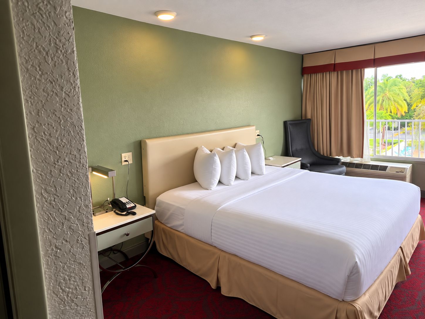 Habitación de hotel con cama, mesita de noche, lámpara y vistas a zonas verdes.