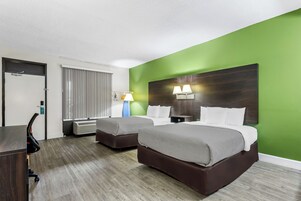 A hotel room with two beds, a desk, a chair, a lamp, and a window with blinds.