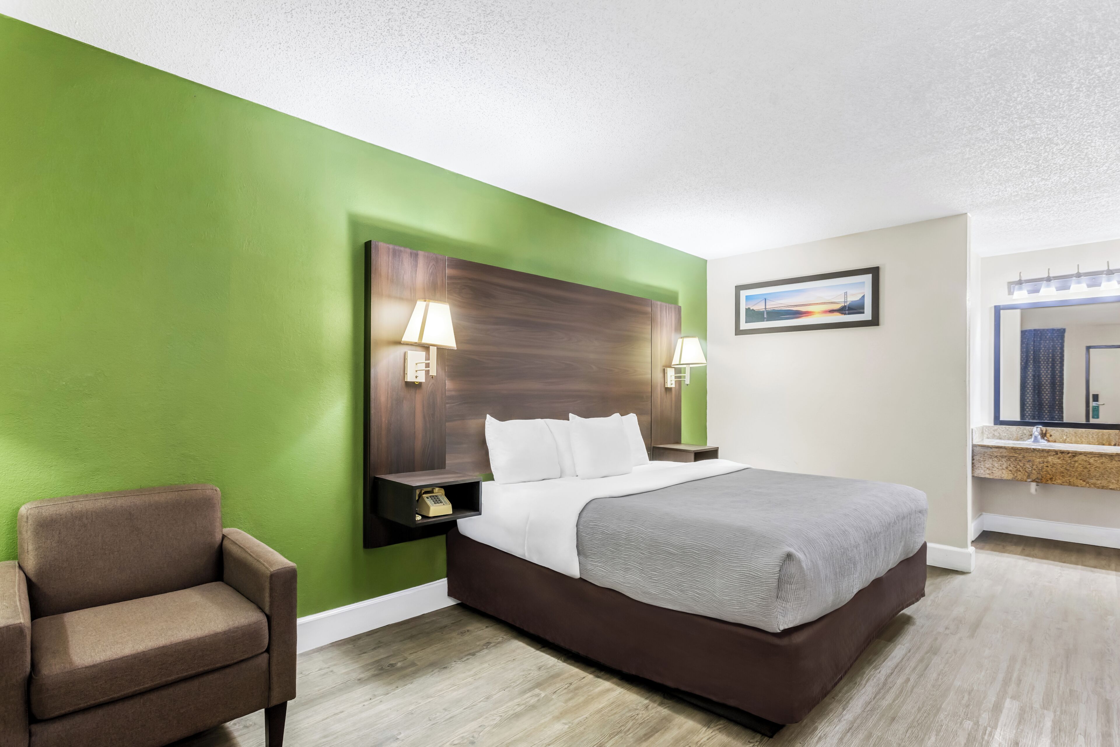 A hotel room with a green wall, a large bed, a nightstand with a lamp, a chair, and a bathroom with a mirror and sink.