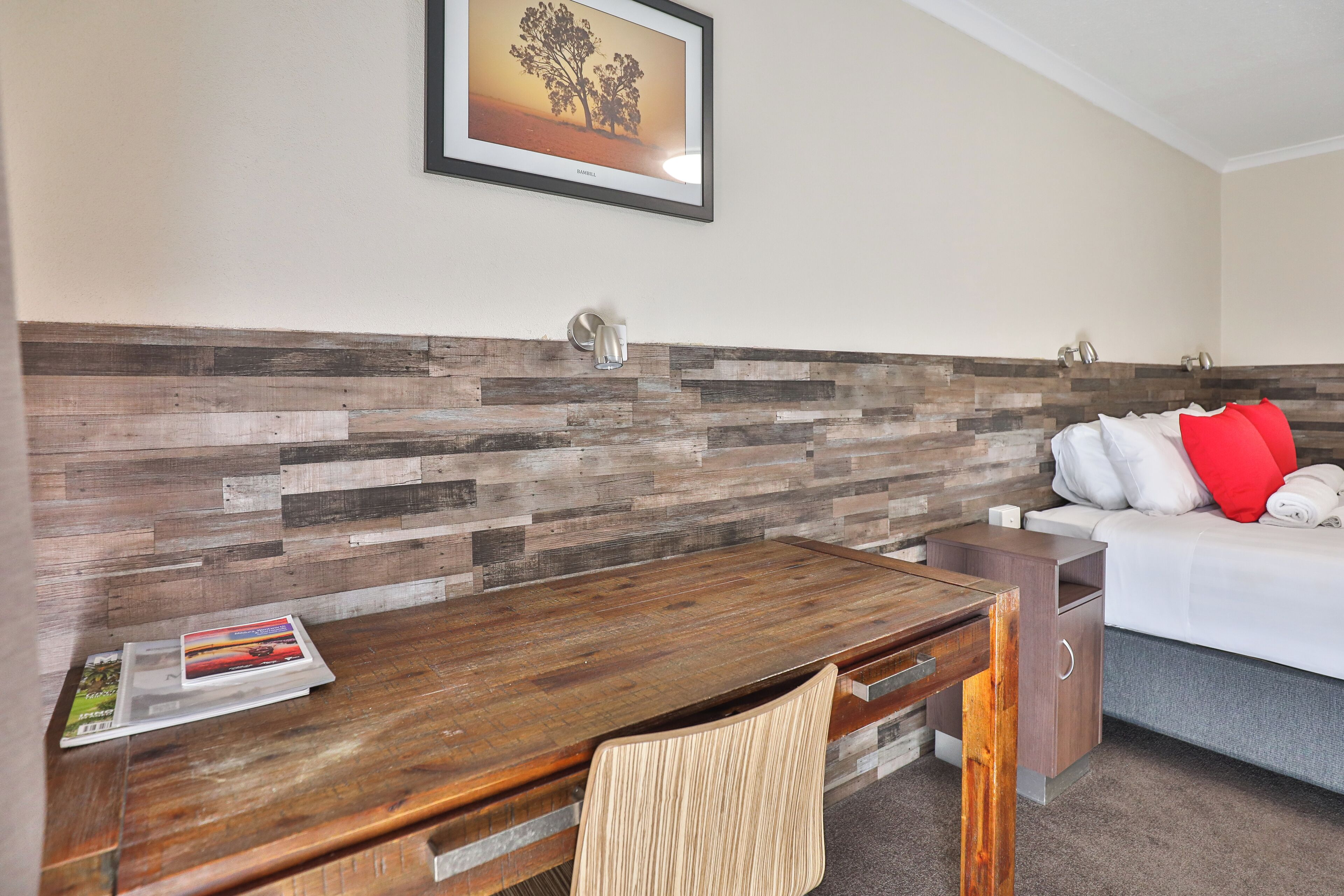 Een hotelkamer met een houten bureau, een bed met witte en rode kussens, en een aan de muur hangend schilderij.