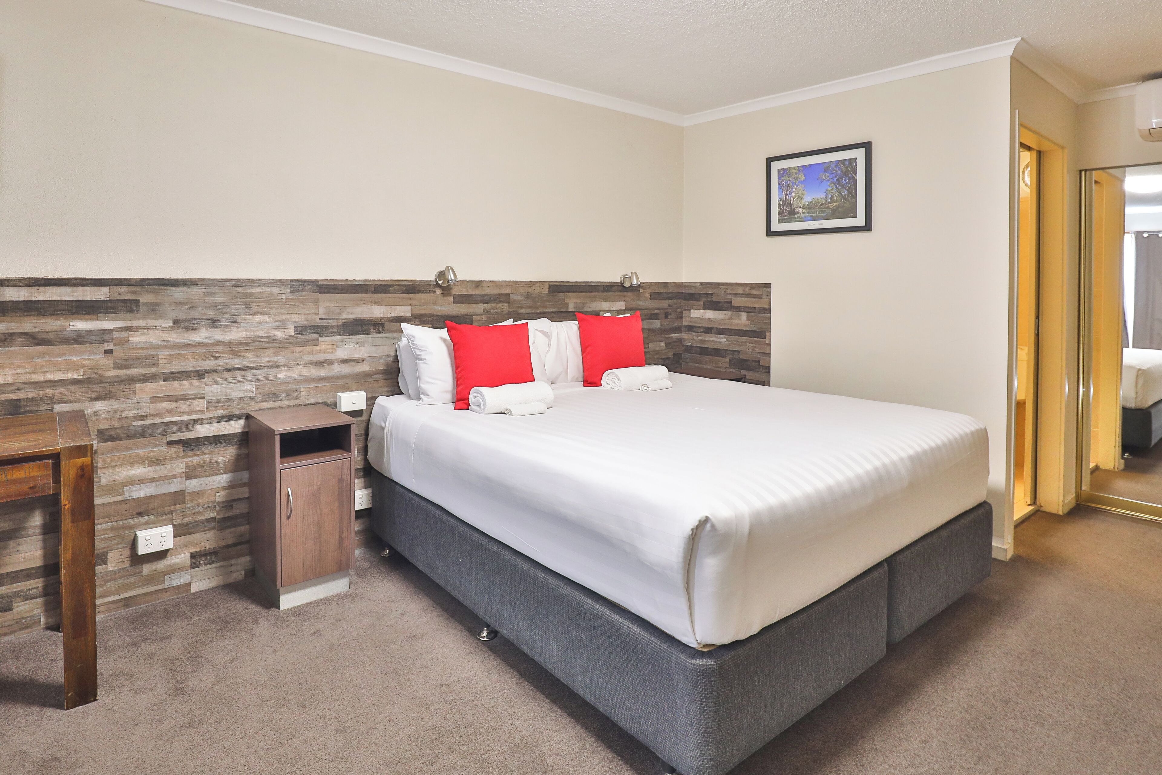 Una habitación de hotel con una cama grande, cabecera de madera, un mueble pequeño de madera y un cuadro en la pared.