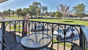 A balcony with a round table and two chairs, overlooking a street with parked cars and a grassy area with trees.