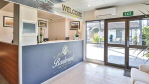 A reception area with a 'Welcome' sign, a reception desk, a 'Boulevard Motor Inn' sign, an 'Exit' sign, and a view of the outside through the glass doors.