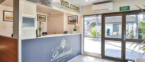 A reception area with a 'Welcome' sign, a reception desk, a 'Boulevard Motor Inn' sign, an 'Exit' sign, and a view of the outside through the glass doors.