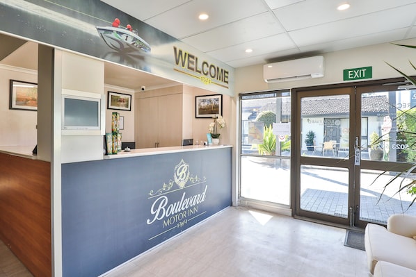 A reception area with a 'Welcome' sign, a reception desk, a 'Boulevard Motor Inn' sign, an 'Exit' sign, and a view of the outside through the glass doors.