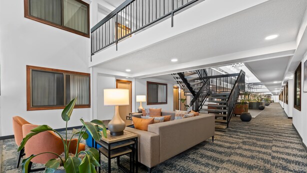 A spacious hotel lobby with a high ceiling, a staircase, a sofa set, and a large potted plant.