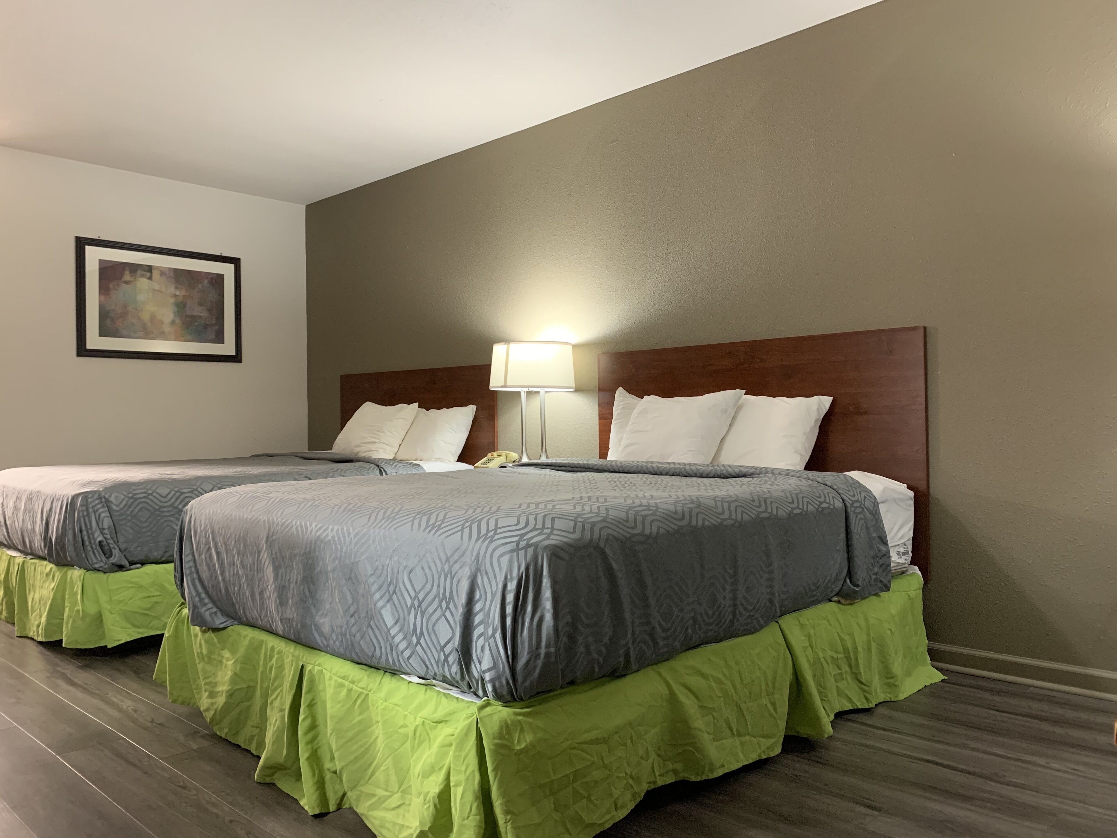 Habitación de hotel con dos camas, cada una con cabecero de madera, ropa de cama estampada en gris y faldas de cama verdes. Se ve una lámpara de noche en una de las camas.