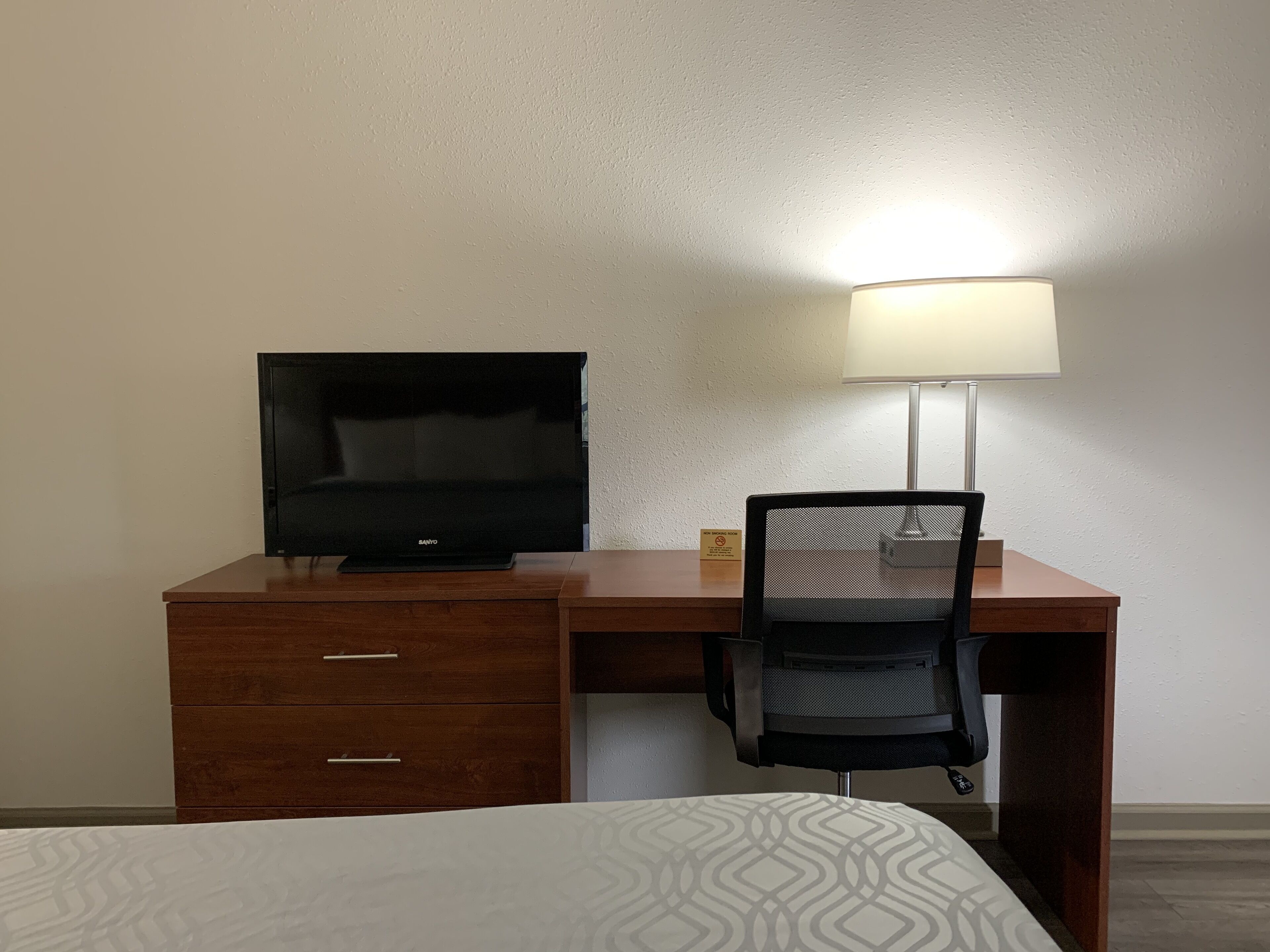 A hotel room with a wooden desk, a black chair, a television, and a lamp.