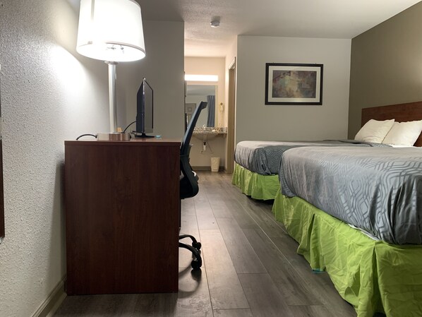 A hotel room with a desk, two beds, a painting on the wall, and a lamp.