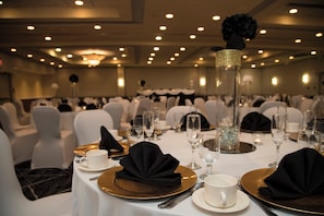Un salón de banquetes con mesas redondas dispuestas para comer, cada una con un plato hondo dorado, una servilleta doblada, una taza blanca y una copa.