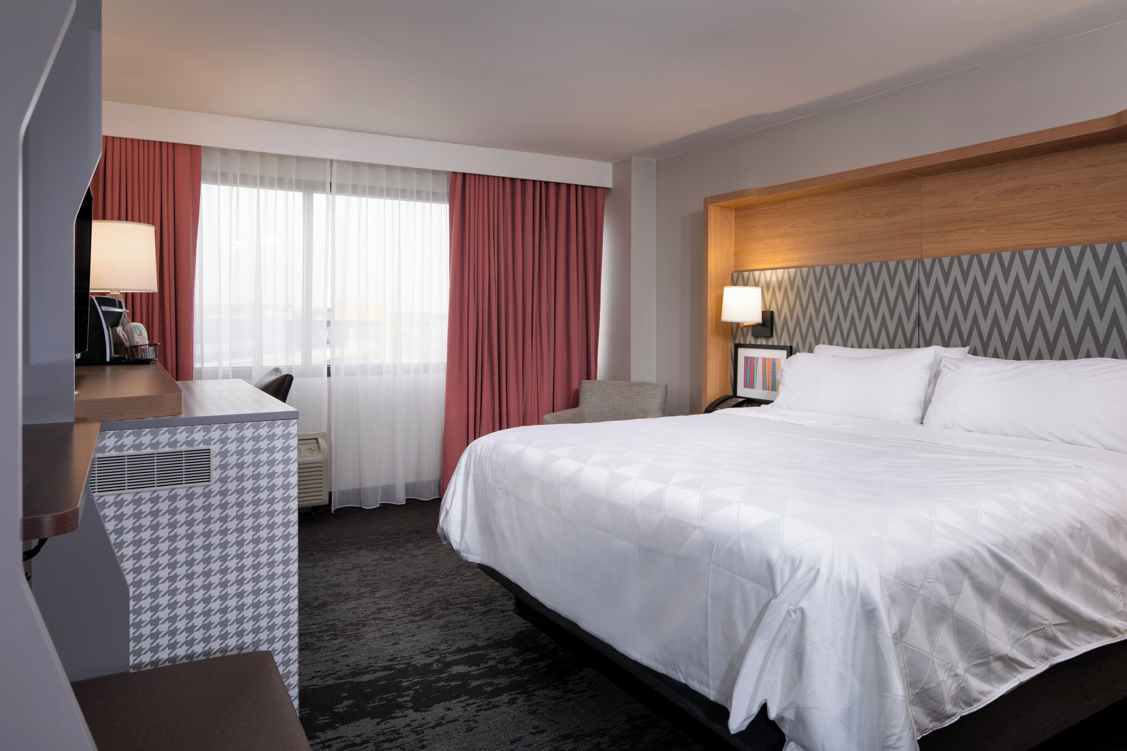 Habitación de hotel con una cama grande, un escritorio con lámpara, una silla y vista a la ciudad desde la ventana.