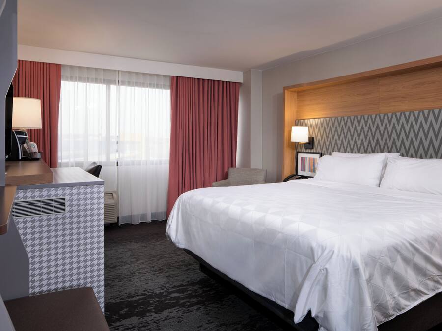 Habitación de hotel con una cama grande, un escritorio con lámpara, una silla y vista a la ciudad desde la ventana.