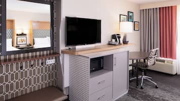 A hotel room with a large flat-screen TV, a desk with a lamp, a chair, a bench, and a large window with curtains.