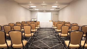 Sala de conferências com fileiras de cadeiras voltadas para uma tela de apresentação, um pódio e piso coberto de carpete.