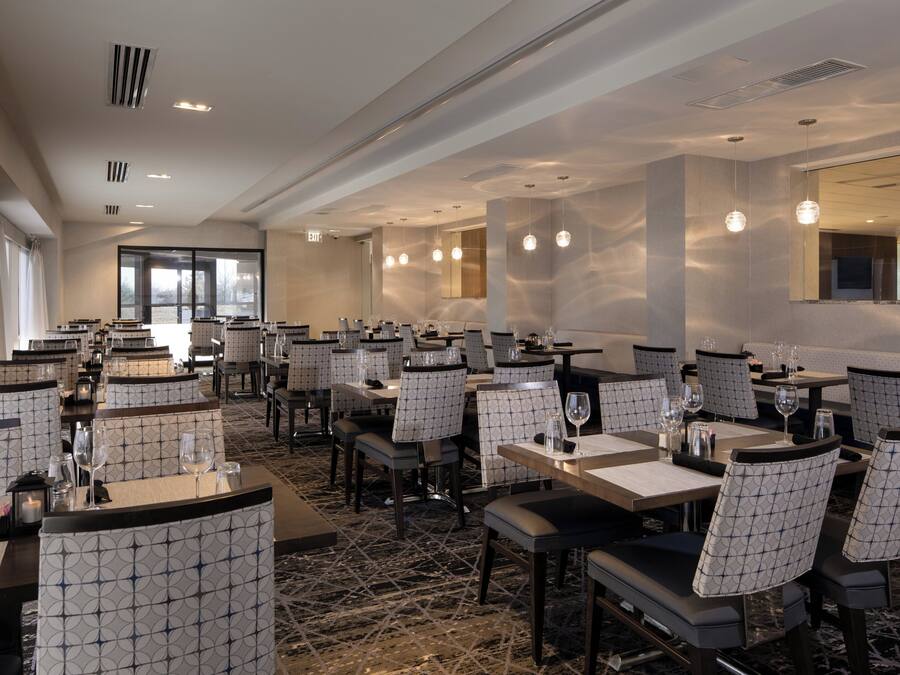 A hotel dining area with multiple tables and chairs, a patterned floor, and a large window providing natural light.