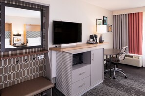 Habitación de hotel con una televisión de pantalla plana grande, un escritorio con lámpara, una silla, un banco y una ventana con cortinas.