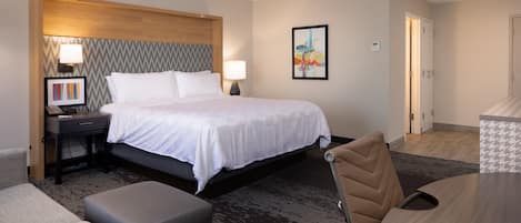 A hotel room with a bed, a desk, a chair, a lamp, and a painting on the wall.