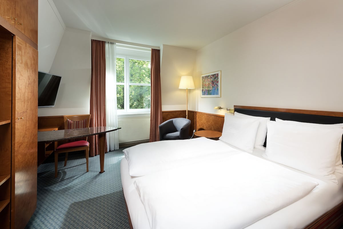 A hotel room with a large bed, a desk with a chair, a TV, a window with curtains, a lamp, and a painting on the wall.