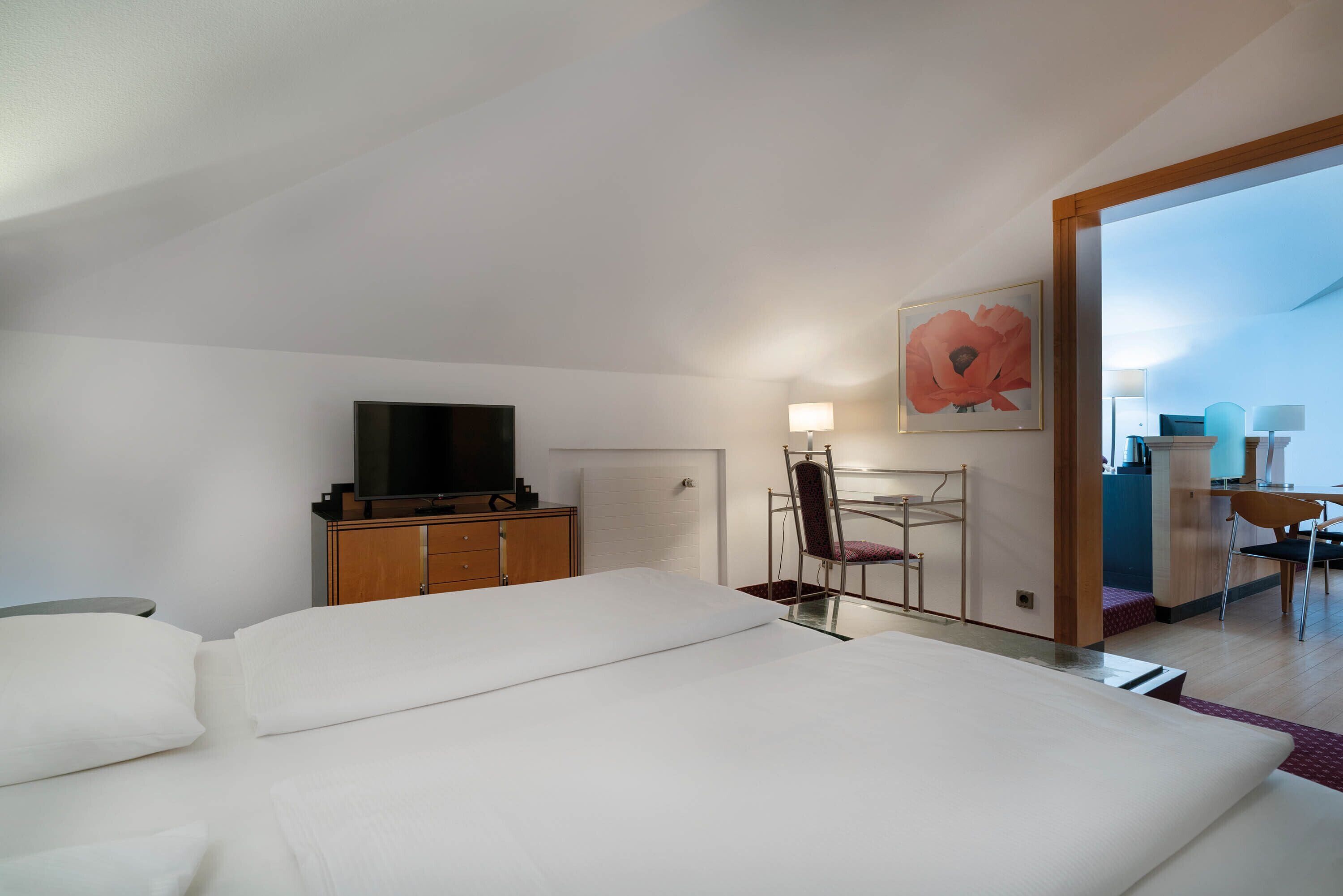 Et moderne hotelværelse med en stor seng, et fjernsyn, et skrivebord med stol, og et stort blomsterbillede på væggen.