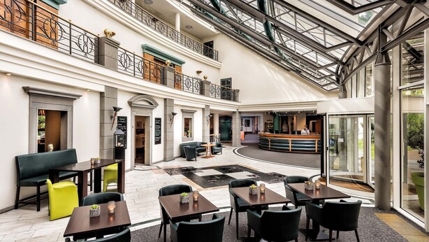 A spacious indoor area with multiple seating arrangements, including tables and chairs, and a central seating area with a curved bench. - Seminaris Hotel Leipzig (Leipzig)