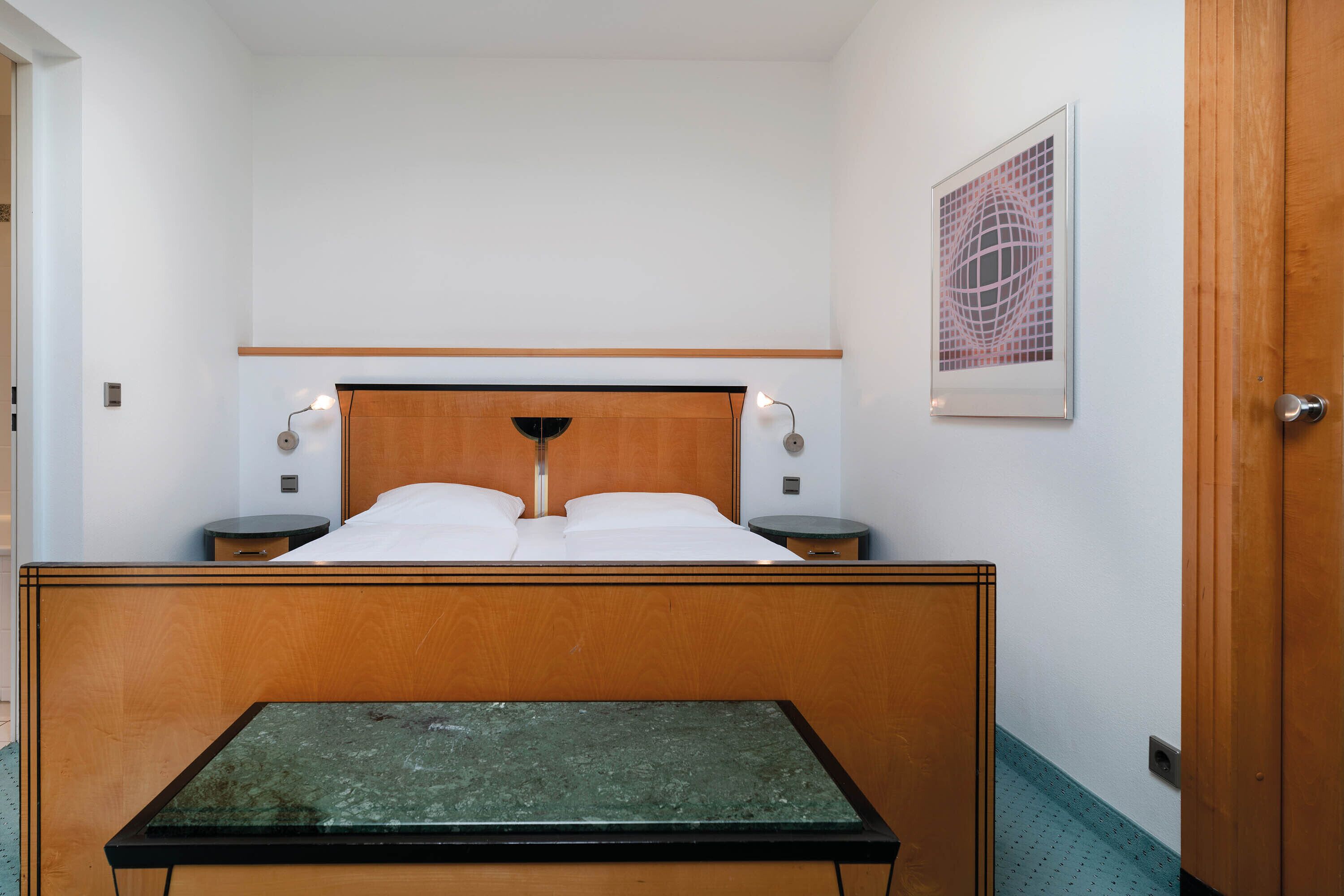 Een hotelkamer met een houten hoofdeinde, een bed met witte lakens, twee nachtkastjes met lampen, een lijst met een kunstwerk aan de muur, en een houten deur.