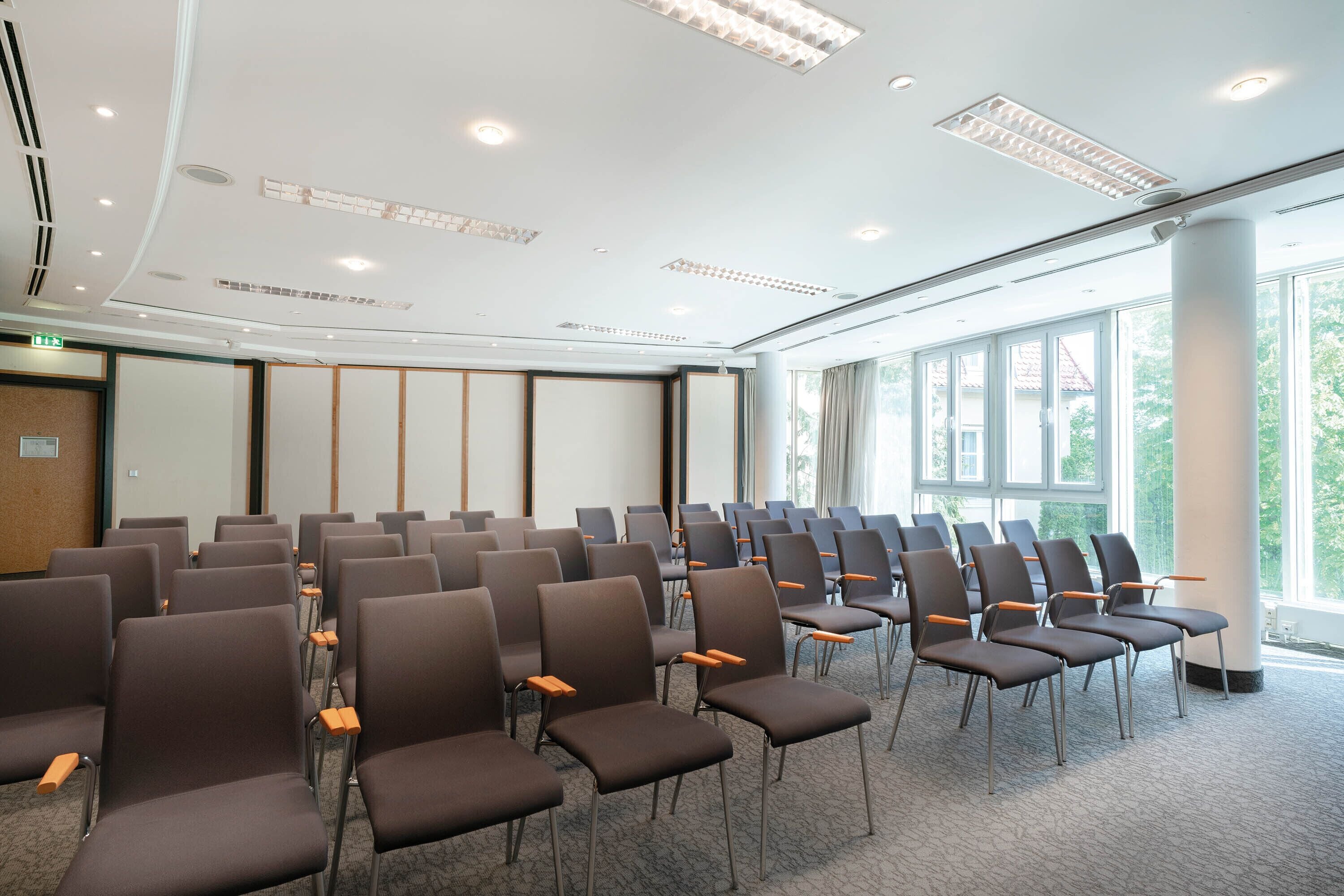 Ein Konferenzraum mit Reihen von Stühlen, einer weißen Decke mit eingelassener Beleuchtung und großen Fenstern, die für natürliches Licht sorgen.
