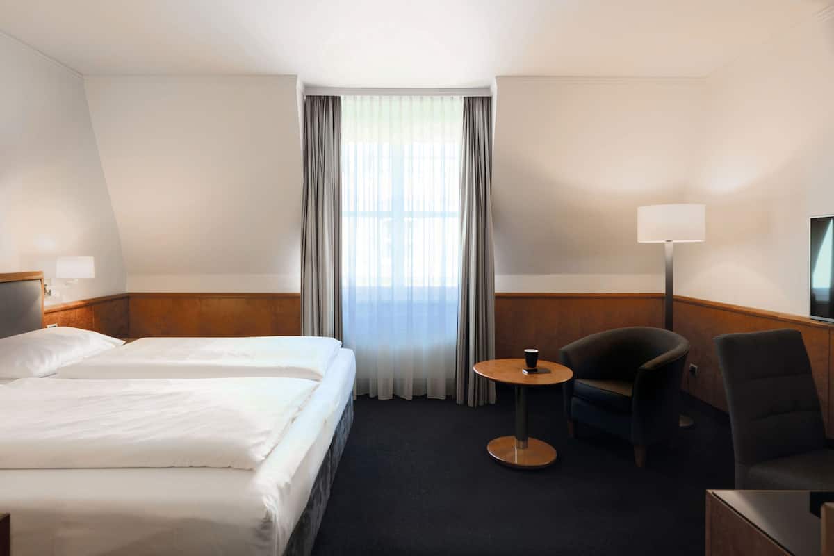 A hotel room with two beds, a small round table, a chair, a lamp, and a TV on the wall.