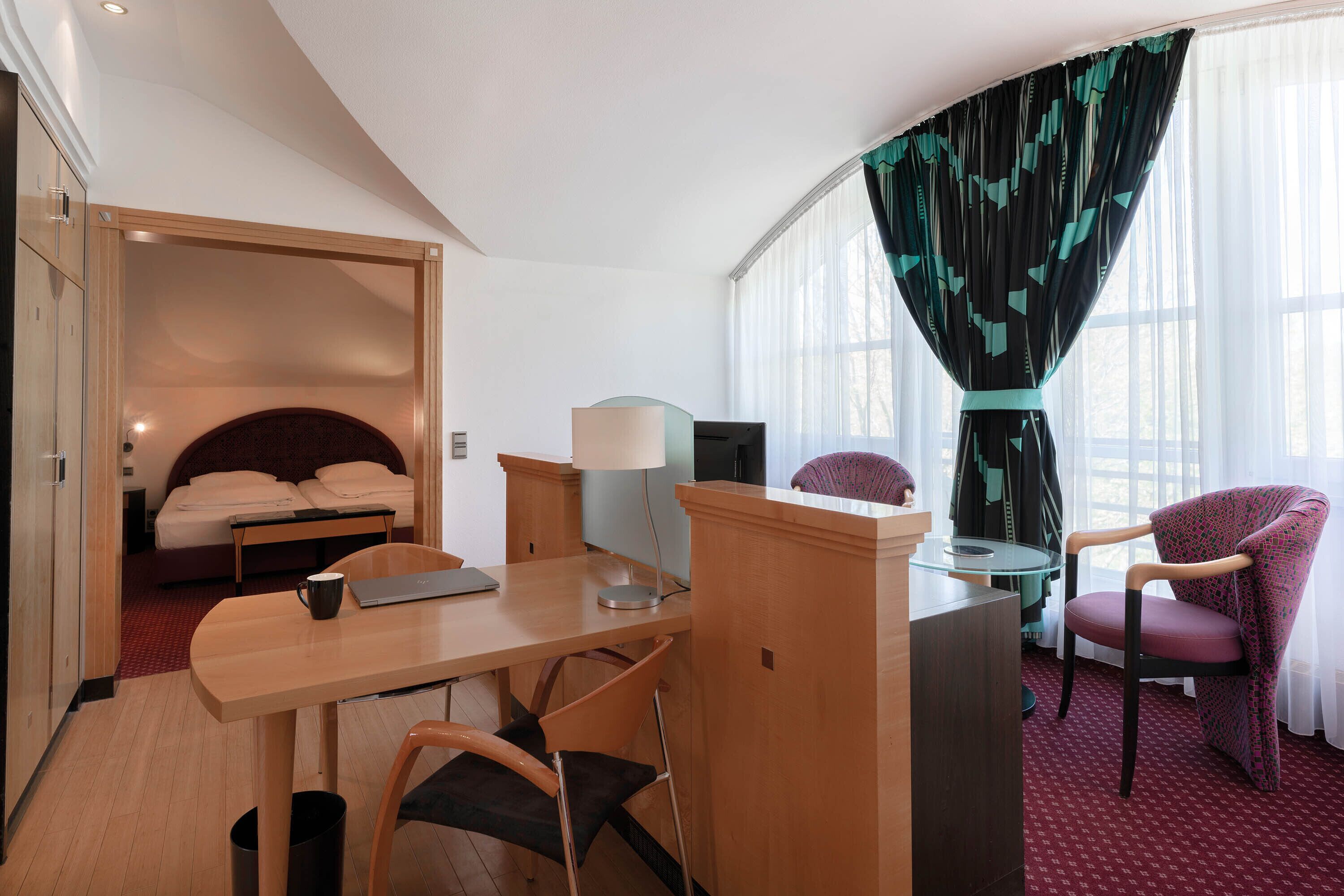 Habitación de hotel con cama, escritorio con una computadora portátil, silla, lámpara y vista al exterior a través de amplias ventanas.