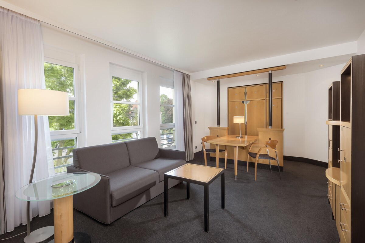 Une chambre d’hôtel moderne avec un canapé gris, une table en verre, une table à manger en bois avec des chaises et une armoire.