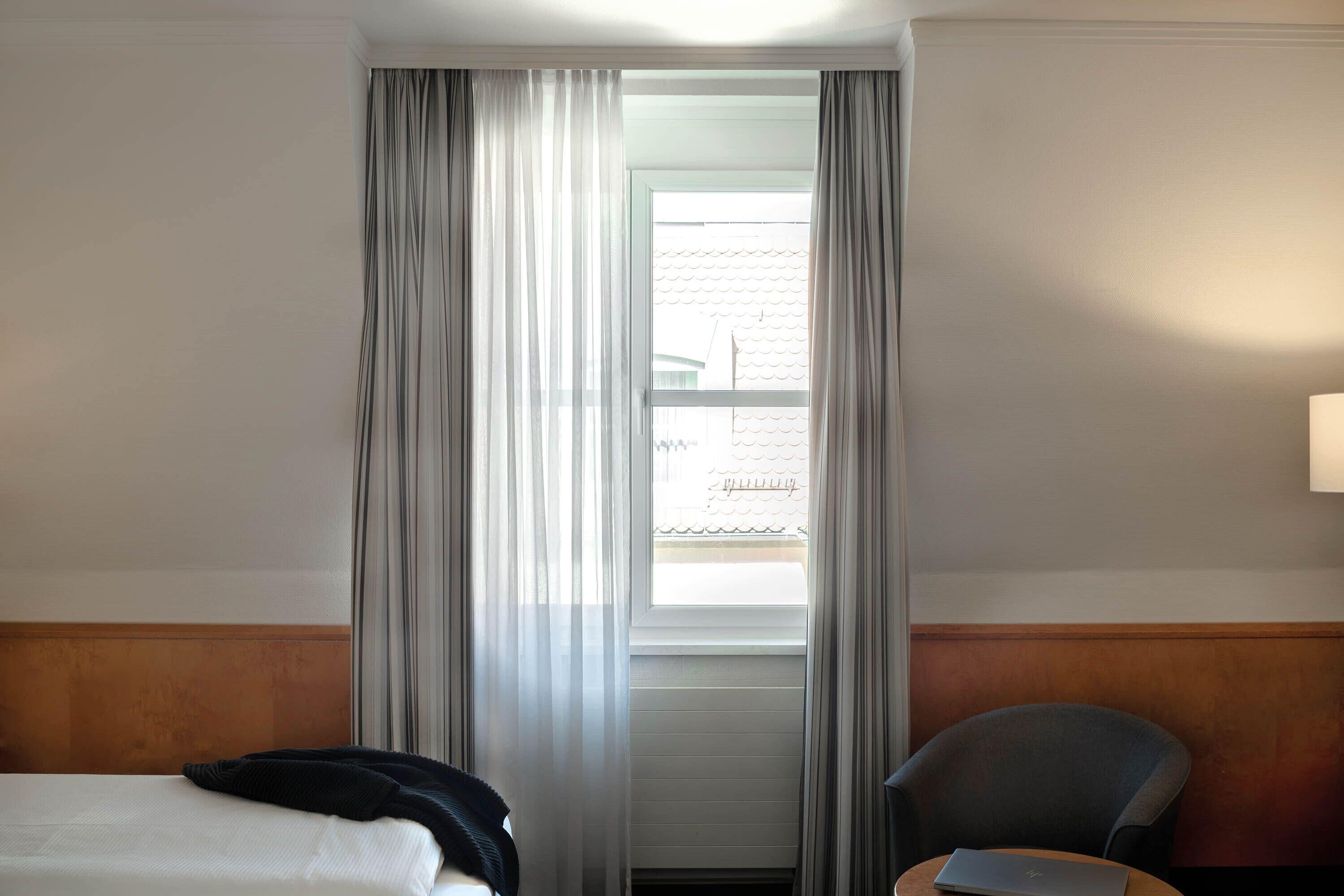 Ein Hotelzimmer mit Bett, Stuhl, einem Laptop auf dem Tisch und einem Fenster mit Vorhängen.